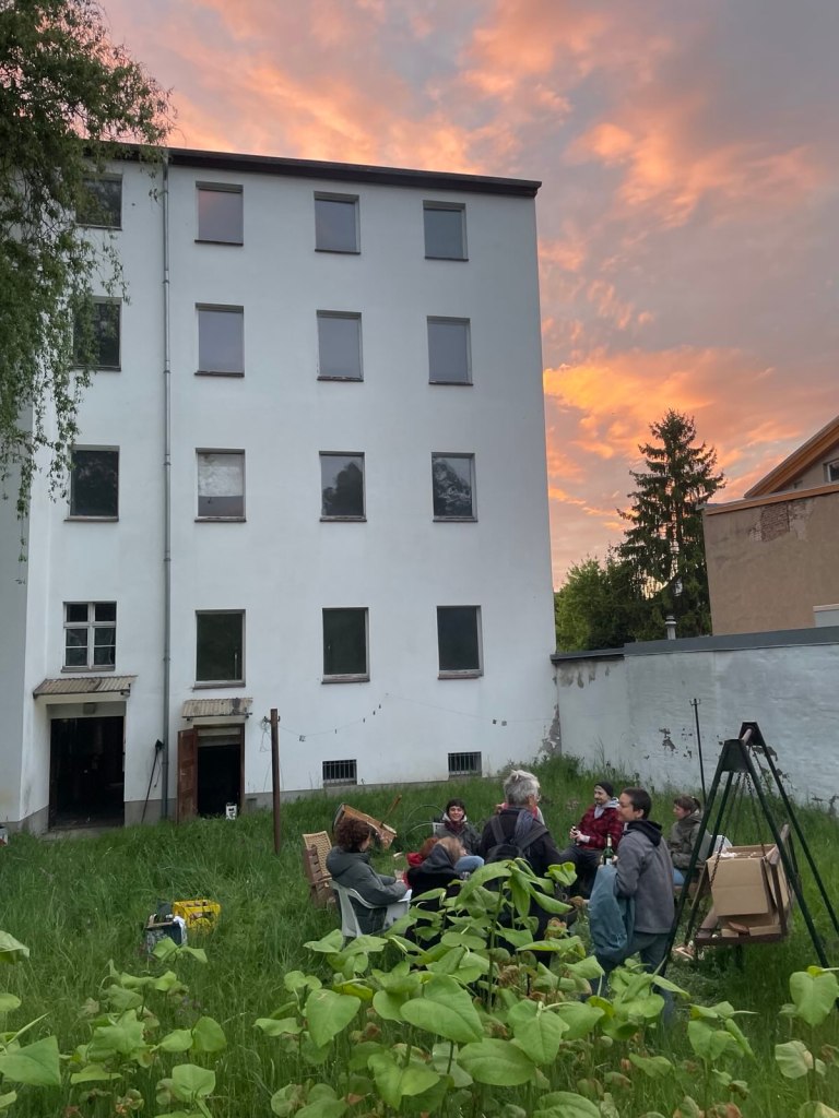 Wir sitzen ums Feuer im Ruh-Garten und plaudern. Im Hintergrund ist das Haus der Ruhlaer Straße 17 in Eberswalde zu sehen. Es ist Abendstimmung mit Sonnenuntergang.